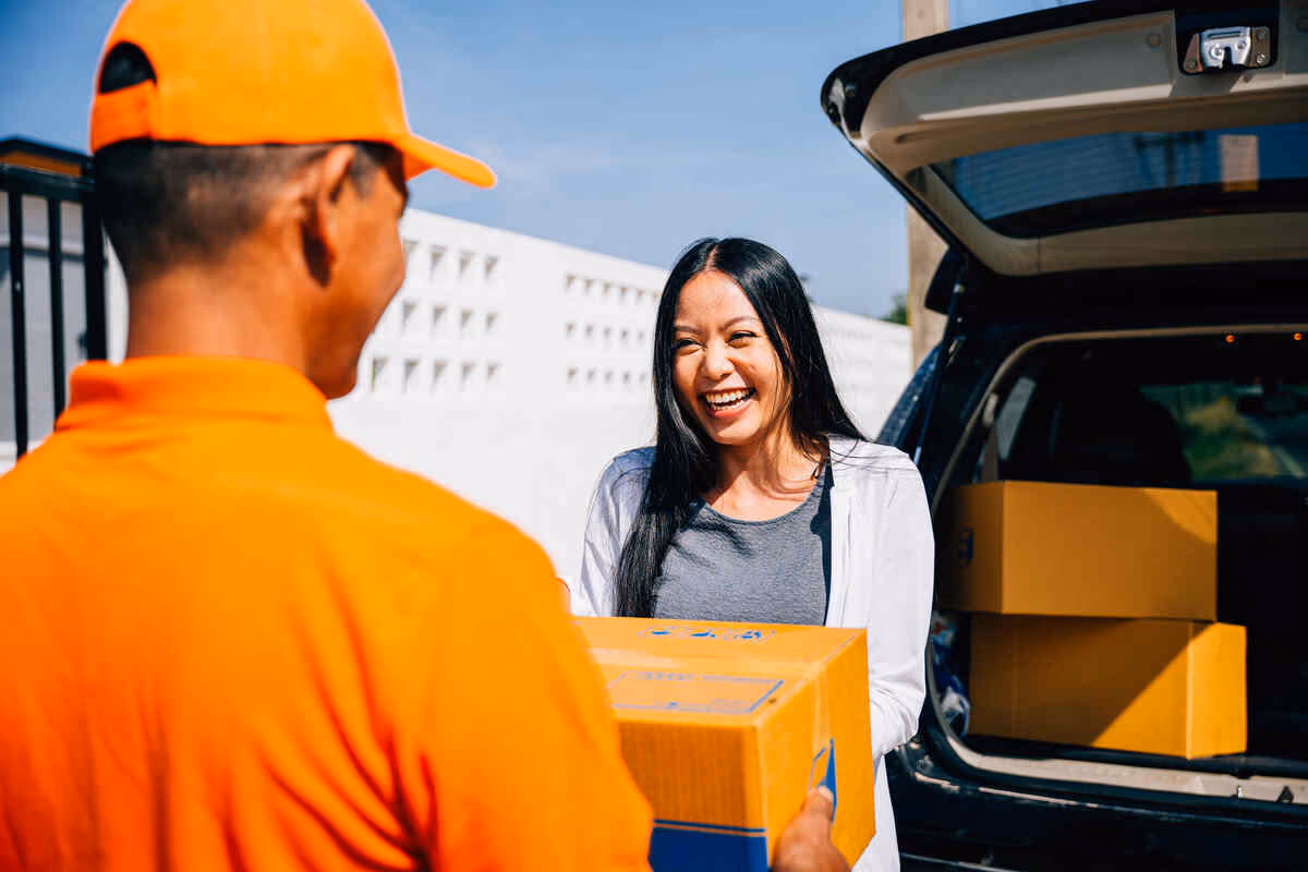 Smiling woman receives a package from a delivery driver beside an open car trunk on a sunny day.