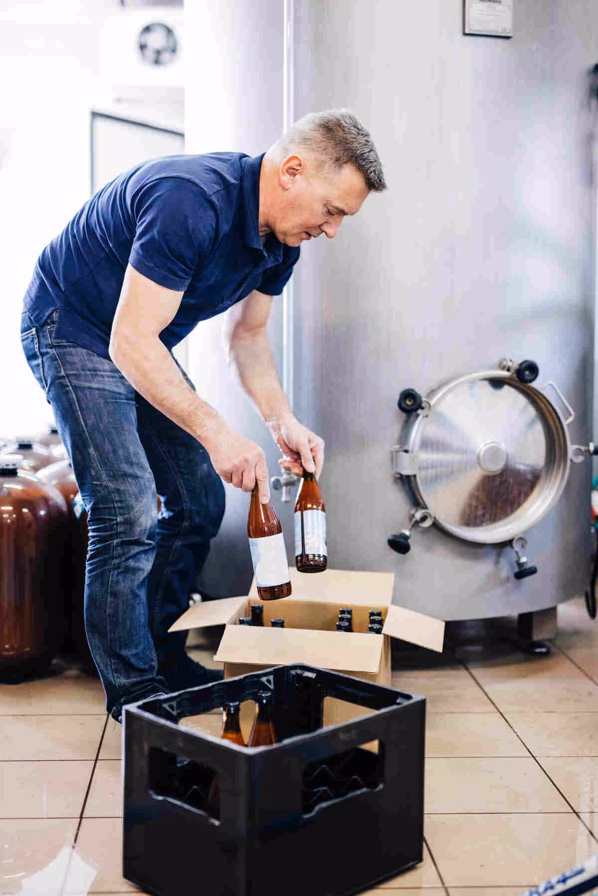 Man in blue shirt and jeans placing beer bottles into a box; metal tank and crate visible.