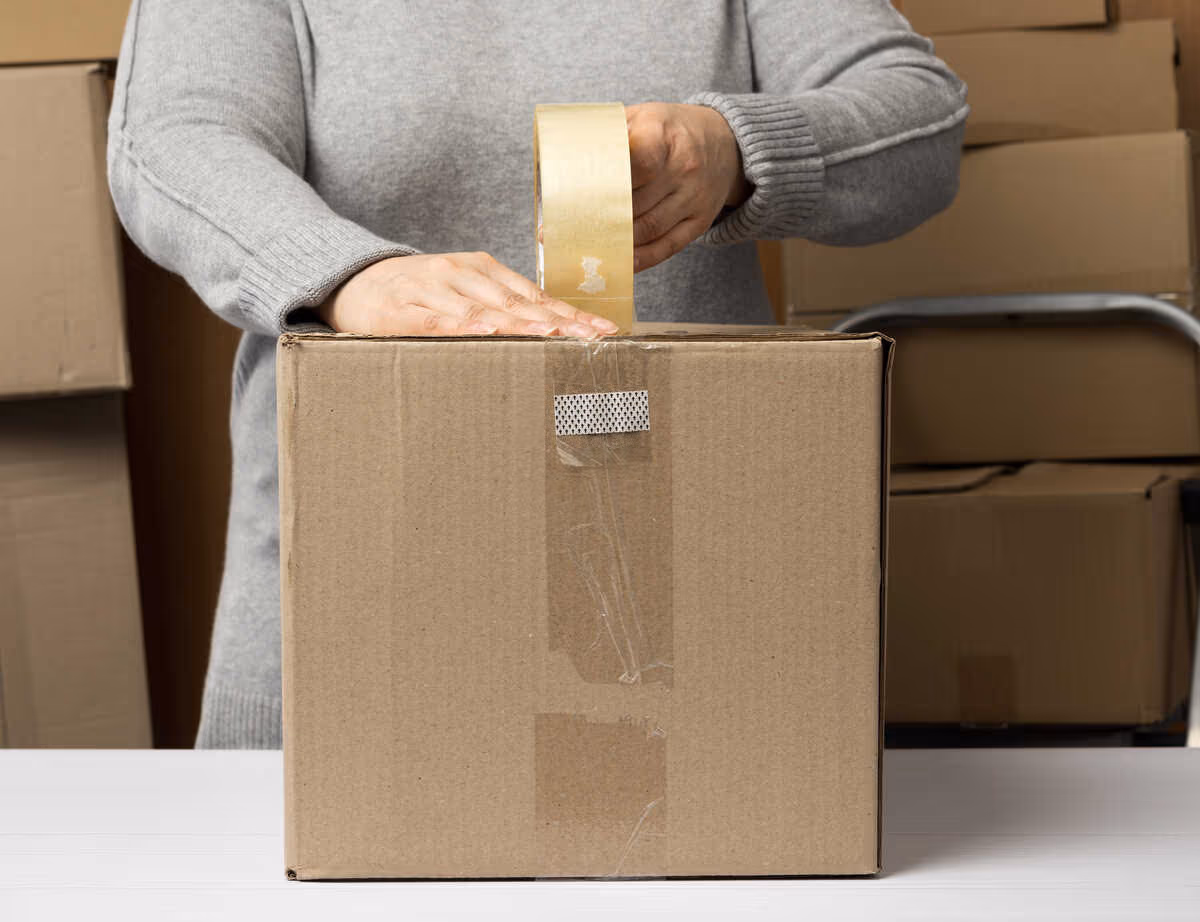 Person seals a cardboard box with packing tape, with stacked boxes in the background.
